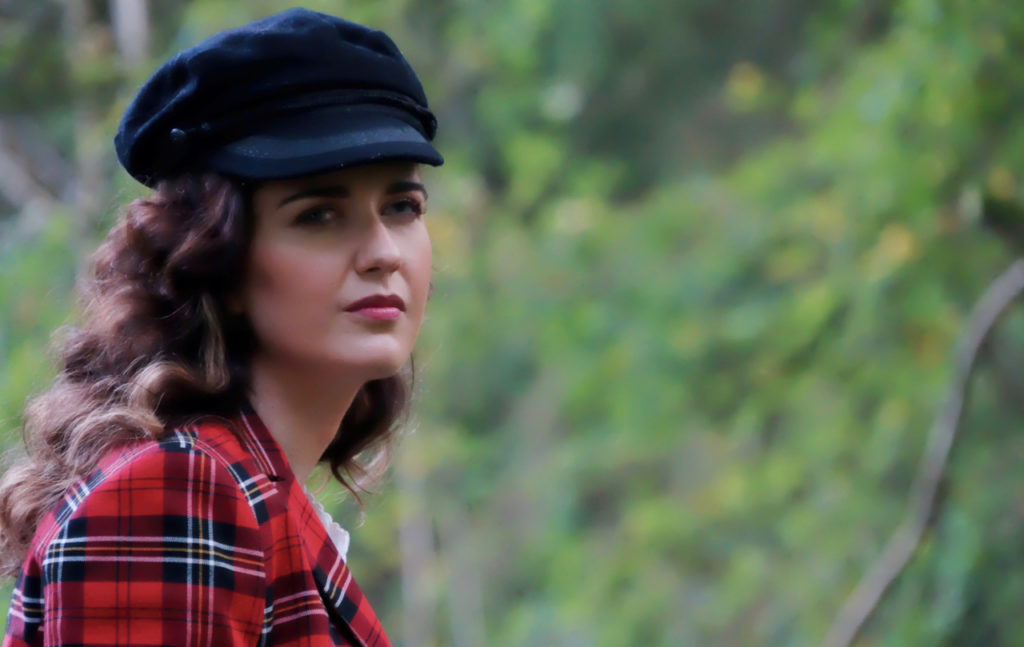 Photo of Andrea in front of some trees, wearing a tartan jacket and peaked cap